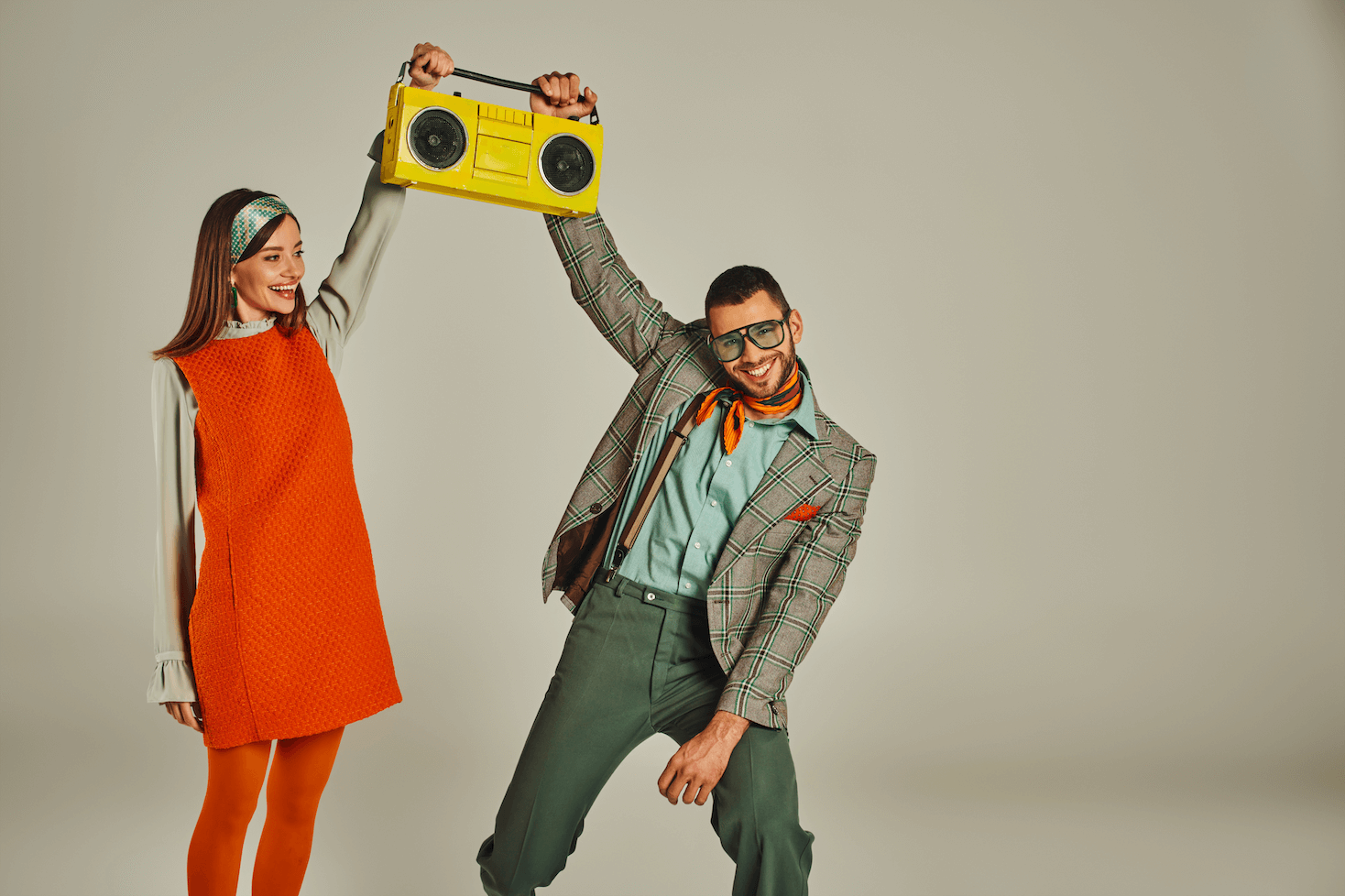 excited couple in stylish vintage clothes holding yellow boombox and dancing on grey backdrop by LightFieldStudios